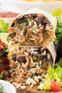 A large, halved burrito stuffed with shredded sweet pork, black beans, and rice, surrounded by lettuce and tomato garnishes.