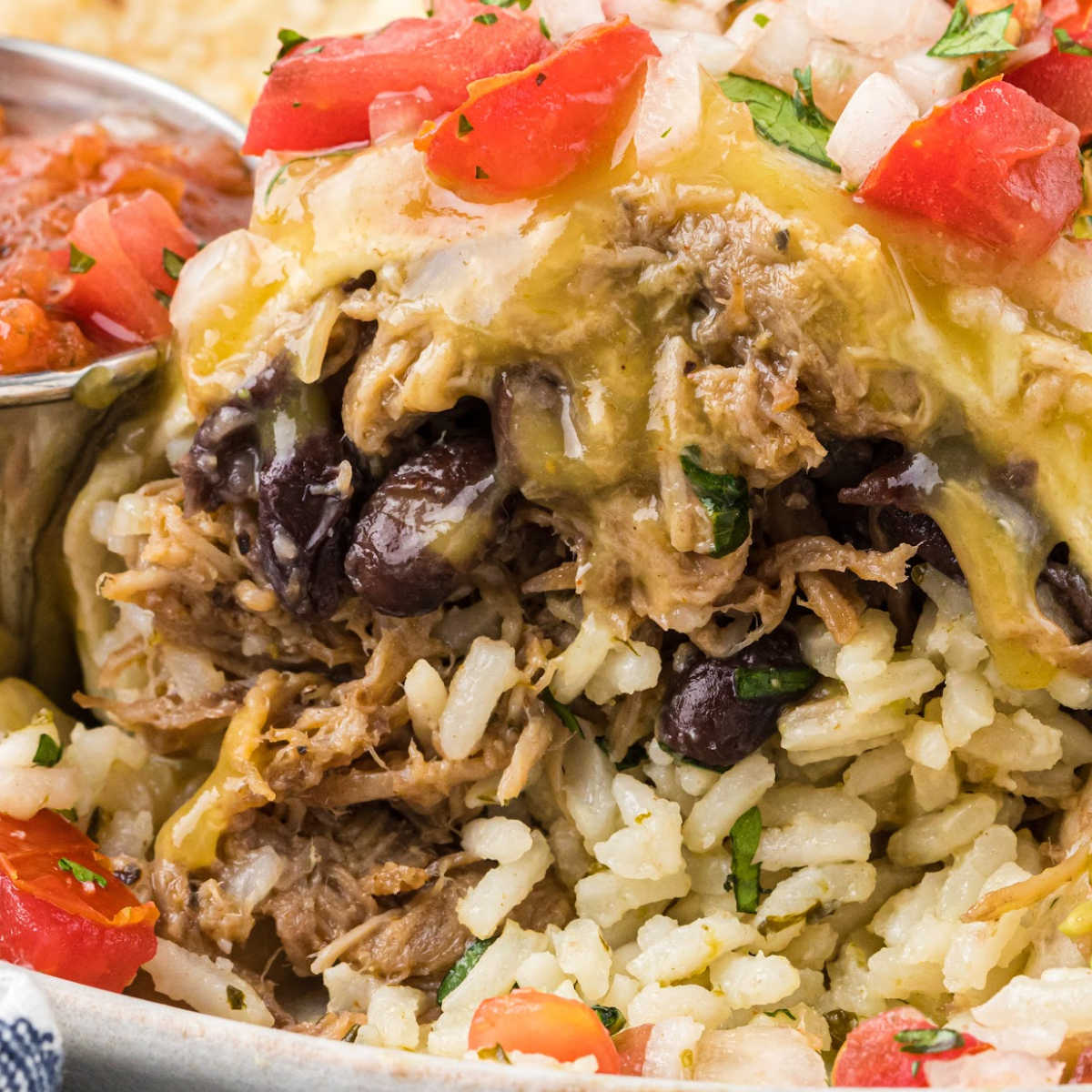 close up of shredded sweet pork in burrito with rice and beans, topped with melted cheese, diced tomatoes, and onions.