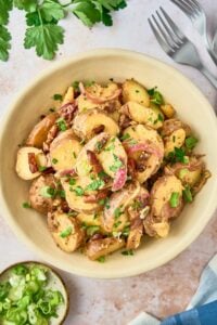 Final dish of Hot German Potato Salad served in a cream-colored bowl, garnished with bacon, green onions, and parsley, with forks on the side.