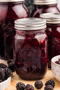 Jars of homemade blackberry pie filling on a wooden table surrounded by fresh blackberries.