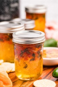 Jars of hot pepper jelly on a wooden surface with crackers and fresh peppers around them.