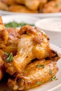 Close-up of golden-brown chicken wings stacked on a plate, with a dipping sauce in the background.