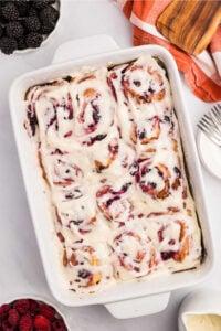 A white baking dish filled with freshly baked sourdough berry sweet rolls covered in creamy white frosting, with a bowl of blackberries and a plate with raspberries nearby.
