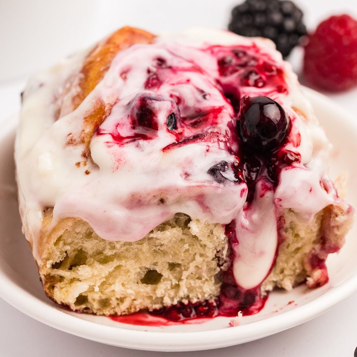A close-up of a sourdough berry sweet roll on a plate, showing the airy texture of the roll and the vibrant berry filling topped with a layer of creamy frosting.