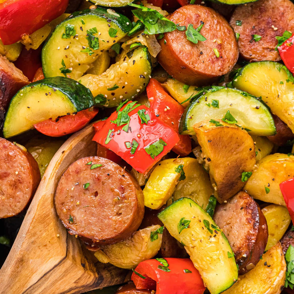 close up of wooden spoon lifting up slices of sausage and veggies topped with parsley.