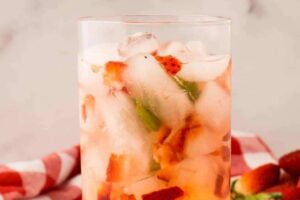 A glass filled with a strawberry shortcake cocktail, packed with ice and garnished with pieces of fresh strawberries and mint leaves.