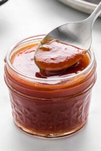 Apricot barbecue sauce in a mason jar, with a spoon lifting some of the sauce, showcasing its smooth texture.
