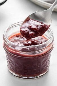 A jar of cherry barbecue sauce with a spoon scooping out some sauce, highlighting its rich, deep red color.