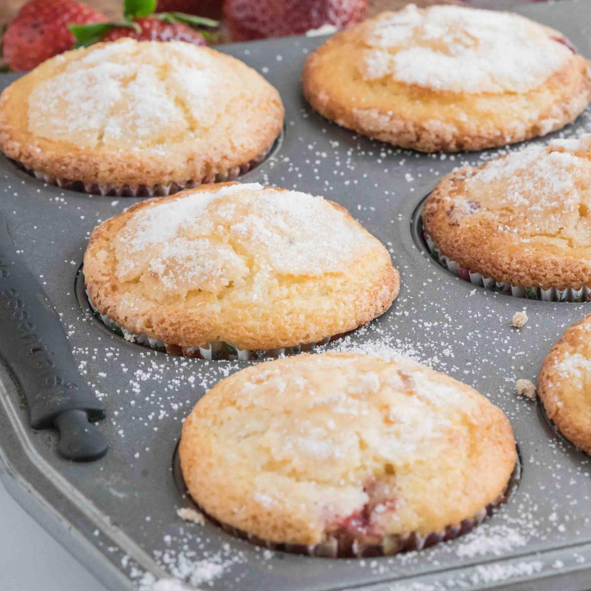 Muffins in a tin, topped with sugar, with a red and white checkered cloth and fresh strawberries nearby.