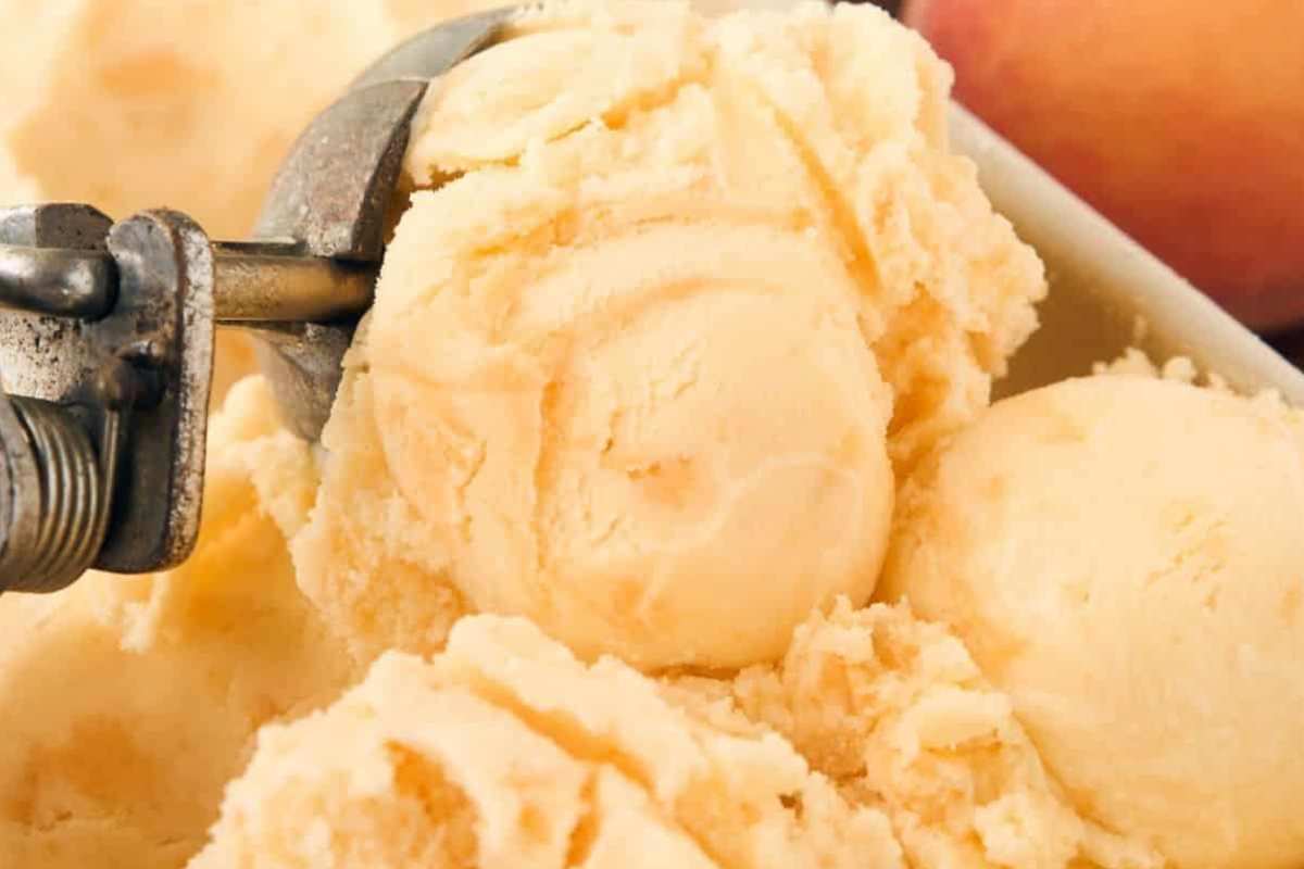 A scoop of fresh peach ice cream, with a peachy hue and chunks of real peaches, being scooped from a container.