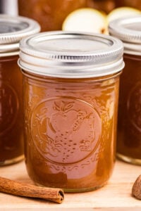 A single mason jar filled with apple pie jam, highlighted with a cinnamon stick nearby and apples in the background.