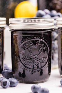 A close-up of a jar filled with blueberry preserves, with blueberries and a lemon in the background.