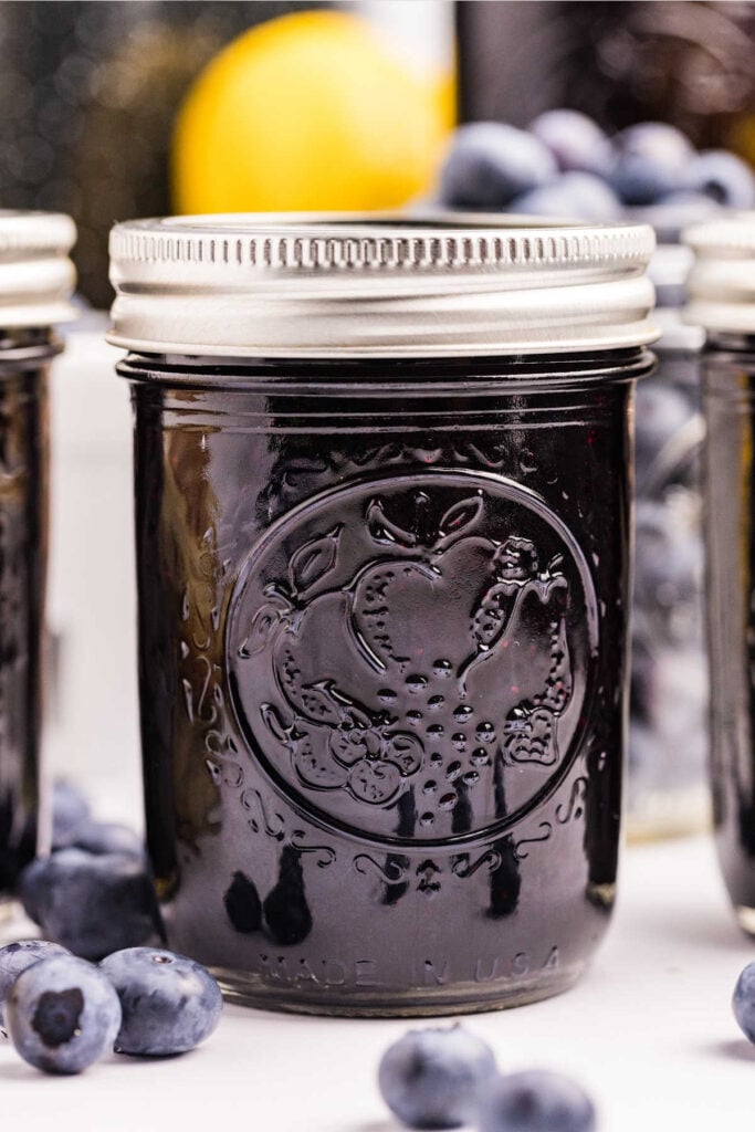A close-up of a jar filled with blueberry preserves, with blueberries and a lemon in the background.
