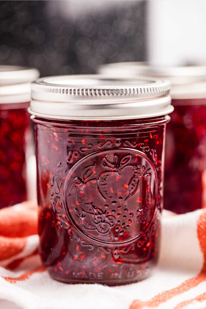 Close-up of a jar of homemade raspberry jam with a detailed lid and an intricate fruit design on the glass. The jar is placed on a striped towel with other jars blurred in the background.