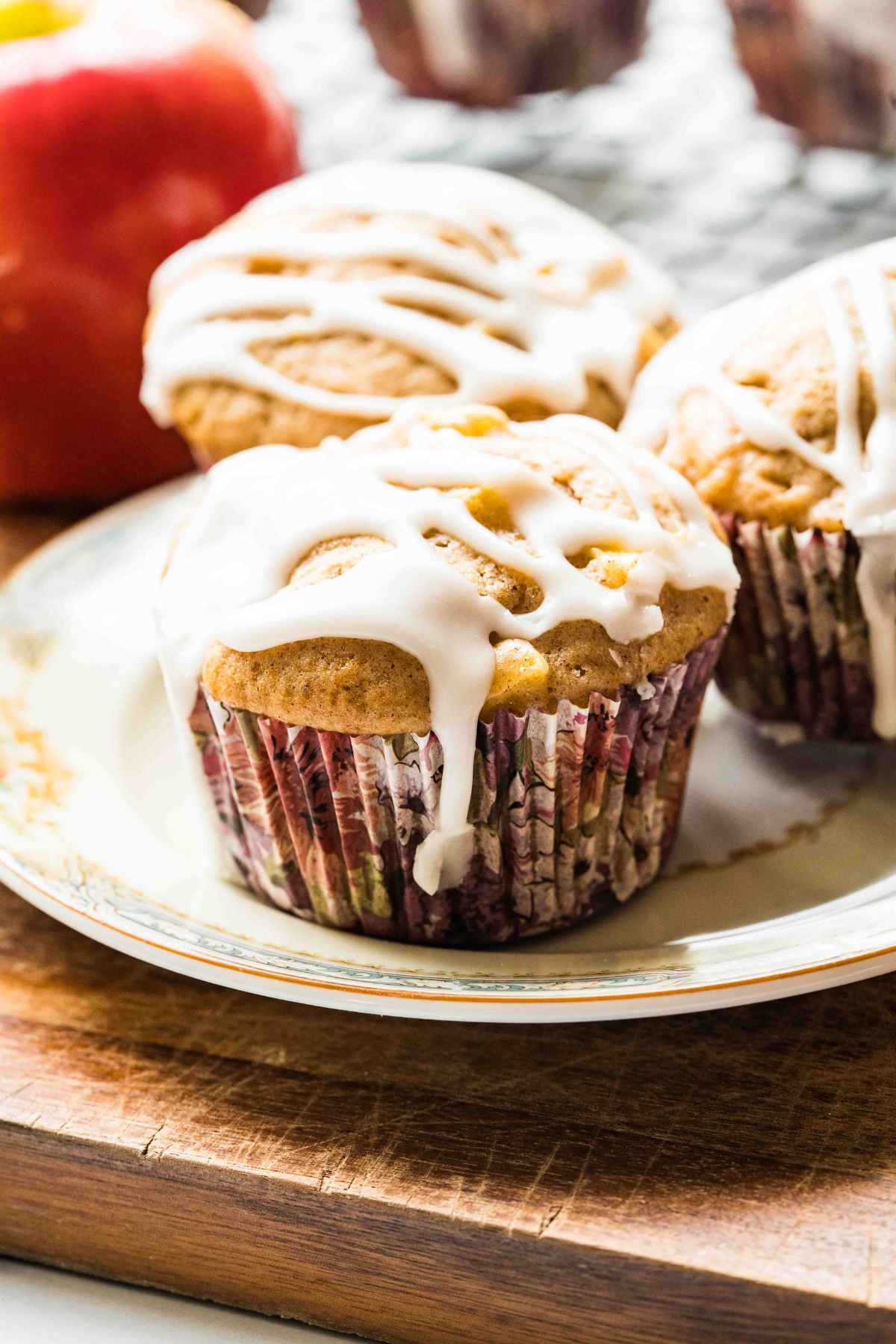 Three sourdough apple cinnamon muffins with white glaze drizzled on top, arranged on a plate with apples and a wooden board in the background.
