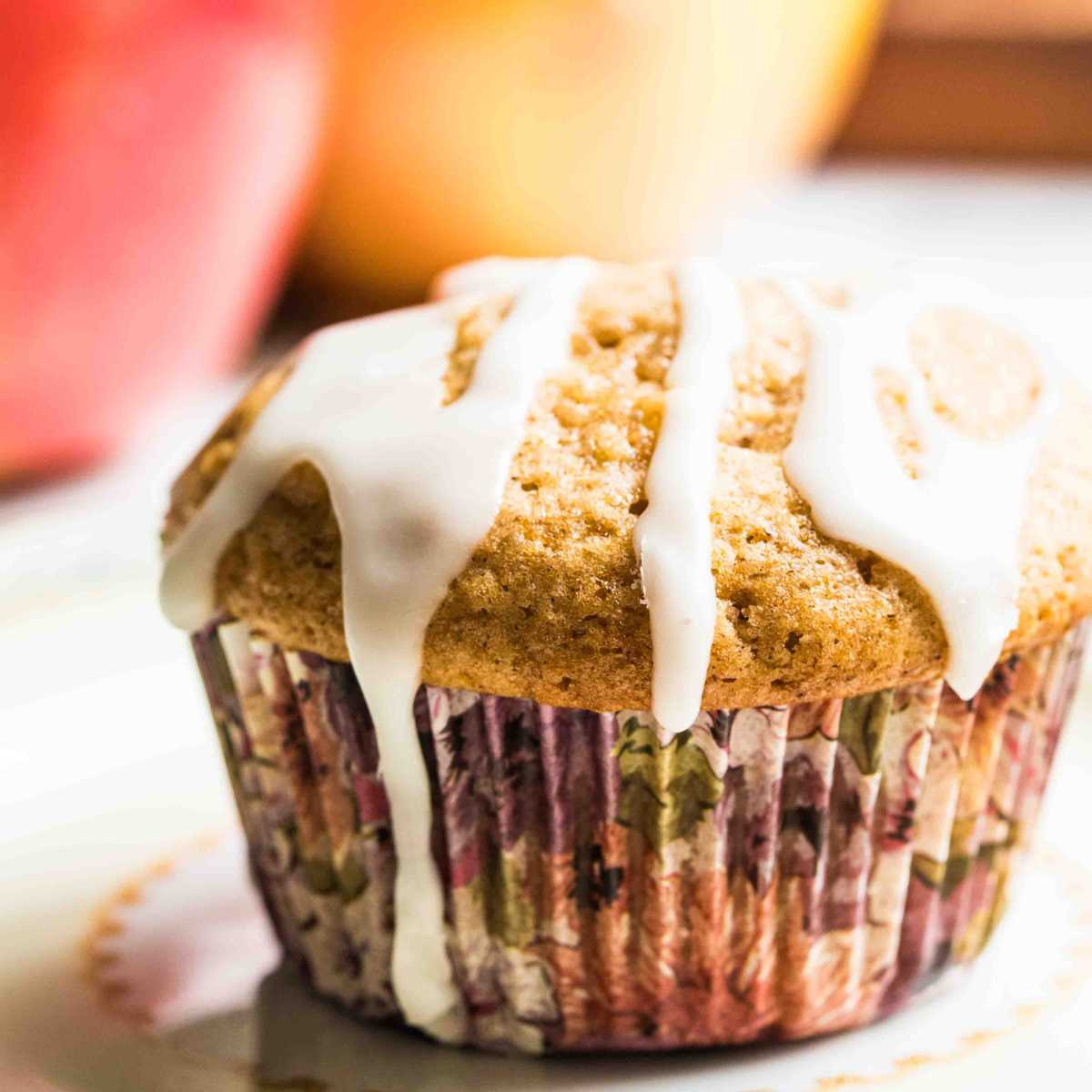 A close-up of a single sourdough apple cinnamon muffin with white glaze drizzled on top, with a blurred background of apples.