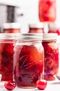 Jars of homemade cherry pie filling with two fresh cherries in front.