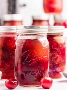 Jars of homemade cherry pie filling with two fresh cherries in front.