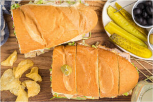 An overhead view of the assembled sandwich cut into pieces, placed on a wooden board, served with potato chips, pickles, and black olives.