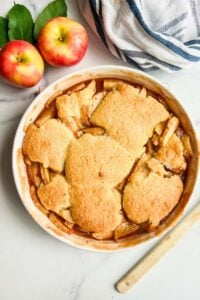 A top-down view of the whole apple cobbler in a round baking dish, with two whole apples and a wooden spoon beside it.