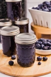 Multiple jars of blueberry syrup on a wooden surface with a bowl of fresh blueberries in the background.