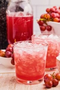 A close-up of two glasses of iced grape juice in front of a jar of freshly canned grape juice, with fresh grapes in the background.
