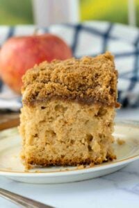 A close-up vertical shot of a slice of Sourdough Apple Spice Coffee Cake on a plate, with a blurred apple in the background.