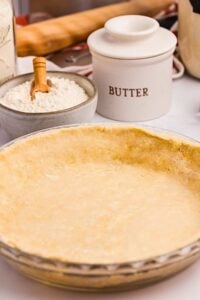 Vertical shot showing the pie crust in a dish with ingredients like flour and butter crock in the background, emphasizing the texture of the crust.