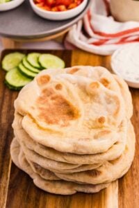 A stack of sourdough pitas on a wooden board with sliced cucumbers and a bowl of tzatziki sauce in the background, ready to serve.