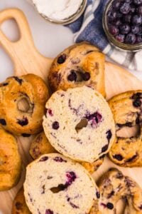An overhead view of blueberry sourdough bagels on a wooden cutting board, with one bagel sliced open to show the soft interior, next to jars of cream cheese and fresh blueberries.