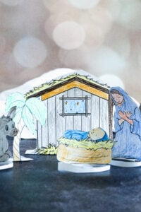 Close-up of a colored and cut-out Nativity scene with baby Jesus, Mary, a donkey, and a stable. The background includes a window with stars and a palm tree.