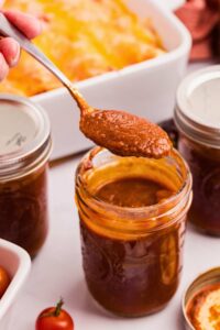 spoon lifting sauce out of jar with pan of enchiladas in the back.