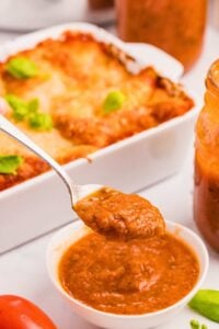 front image of spaghetti sauce in bowl with spoon lifting some out, with dish of lasagna in background.