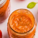 overhead shot of open jar showing spaghetti sauce inside with a closed jar in the top left corner and a small leaf in the top right corner.