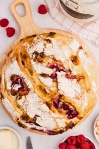 overhead shot of white chocolate raspberry sourdough bread on wooden board with raspberries around it on the counter.