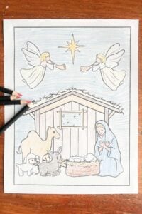 A colored-in Nativity Scene coloring page featuring Mary, baby Jesus, angels, and animals, shown with colored pencils on a wooden table.