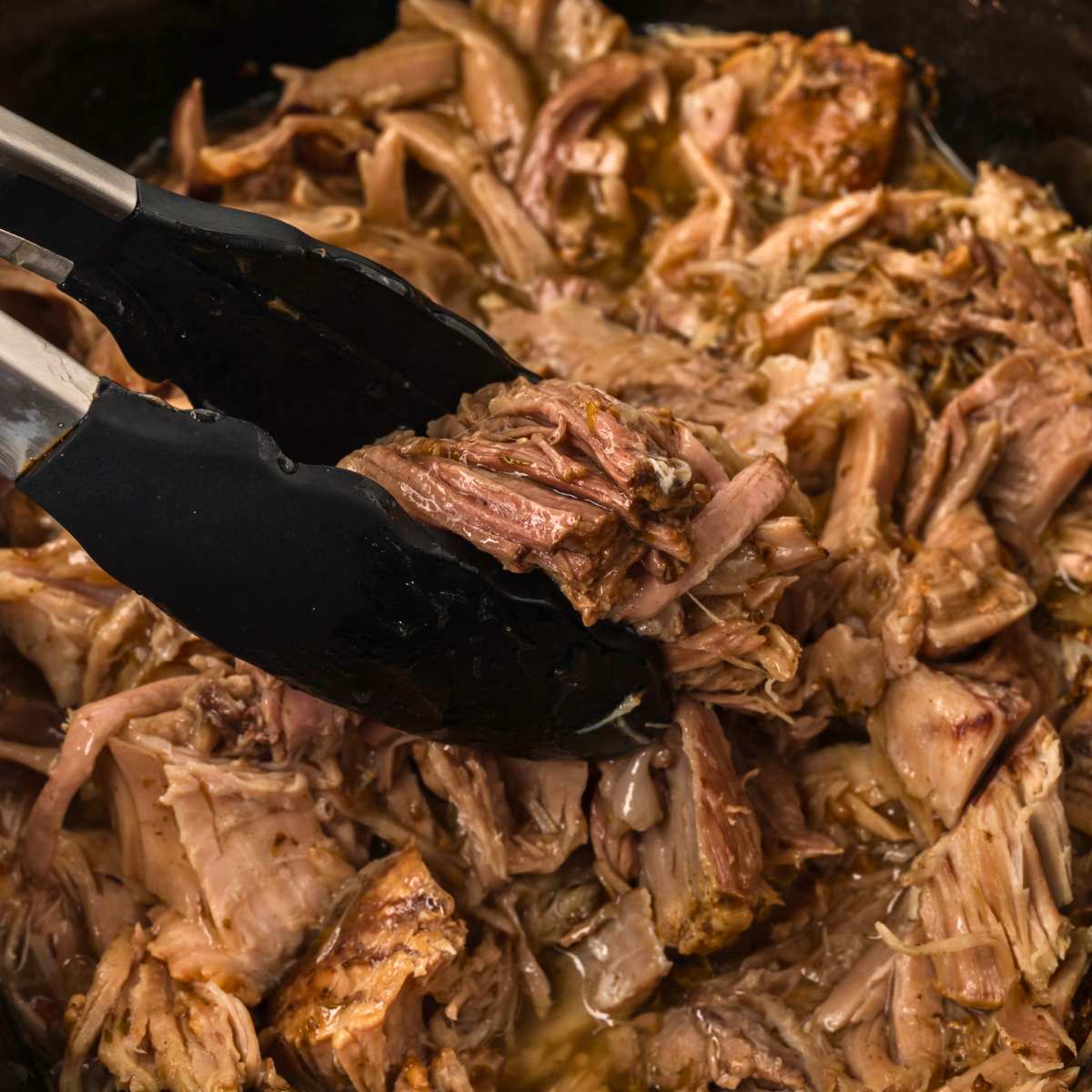 tongs grabbing shredded pork roast with gravy in slow cooker.
