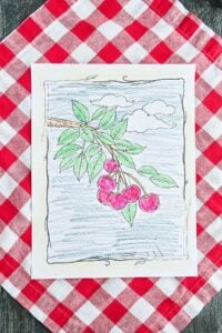 A finished cherry branch coloring page with red cherries and green leaves sits on a red and white picnic tablecloth.
