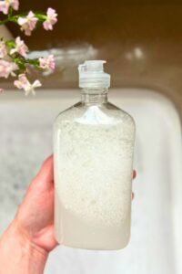 hand holding clear bottle of liquid dish soap over bubbly sink.