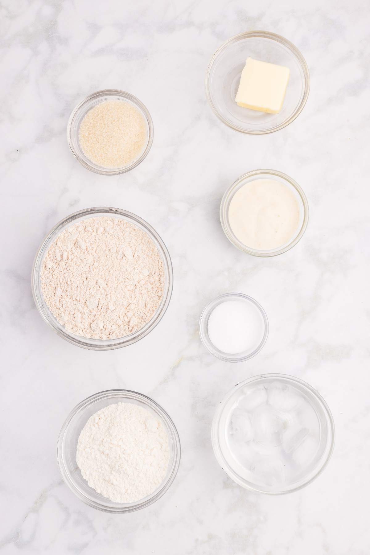 Bowls of flour, sugar, butter, salt, starter, and ice water on a counter.