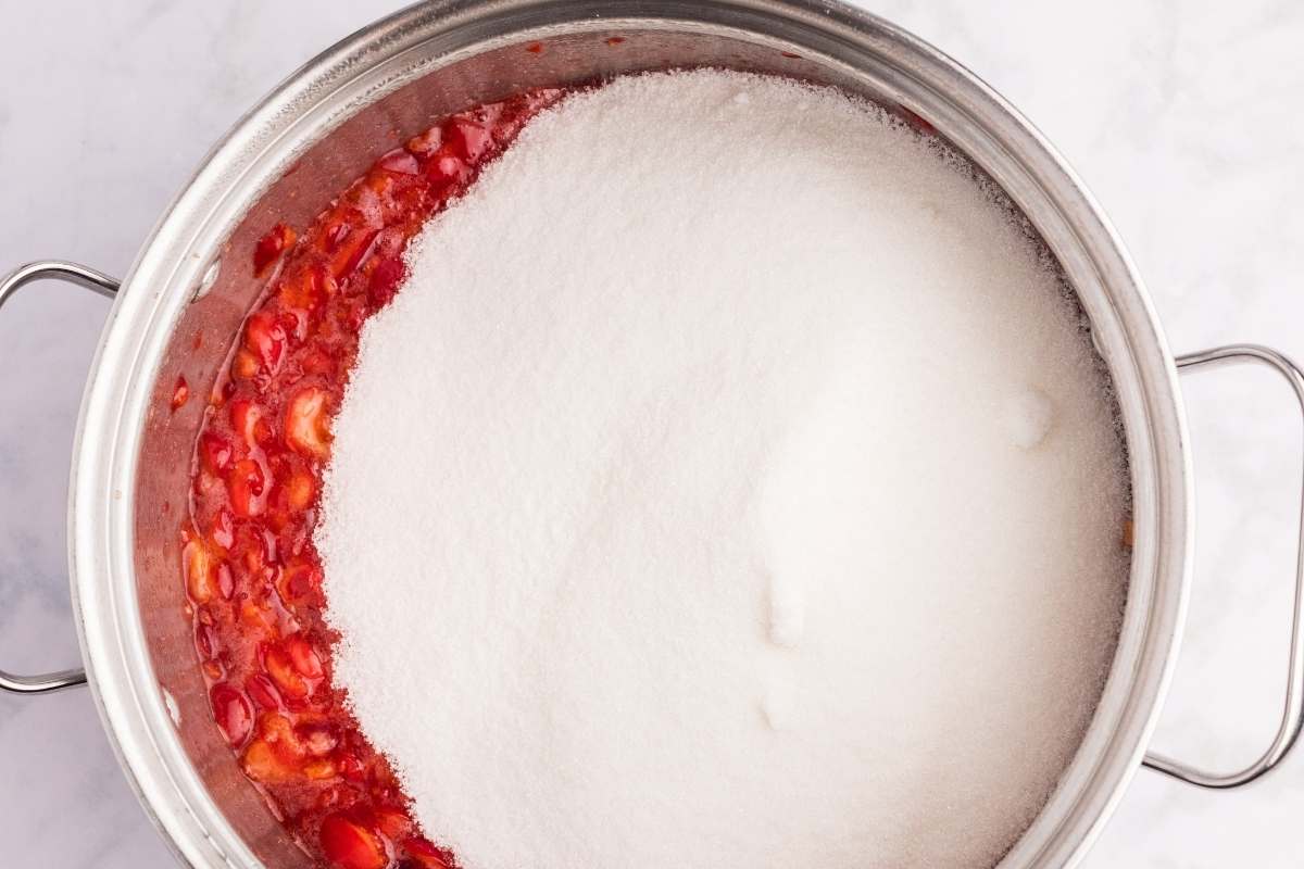 Cherry jam mixture with sugar being added to the pot.