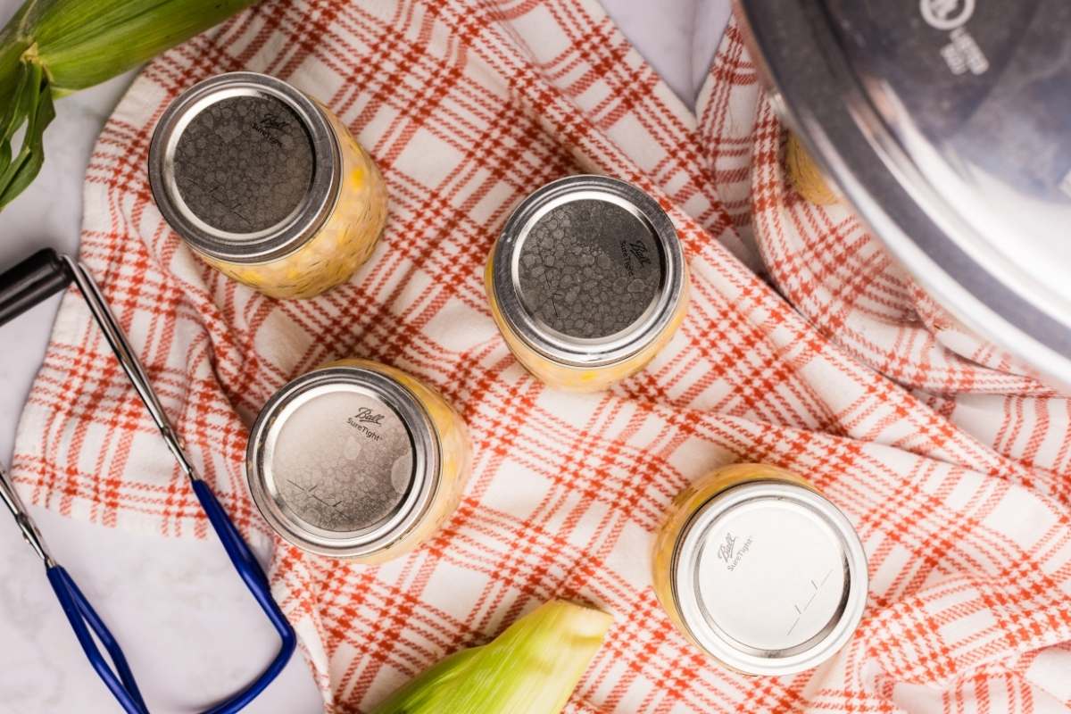 Three jars of corn next to corn cobs and a red towel.