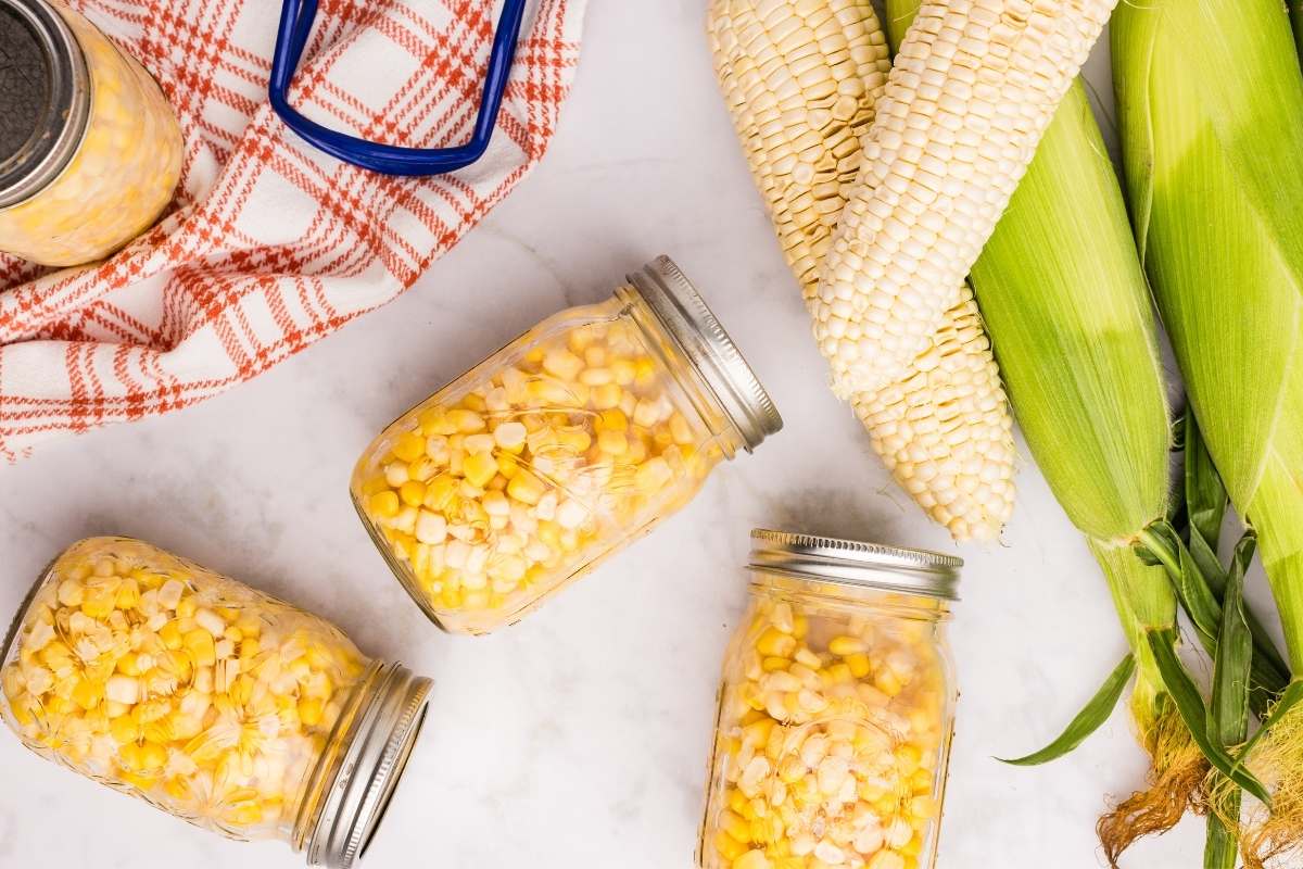 Four sealed jars of corn on a red plaid towel with canning tools.