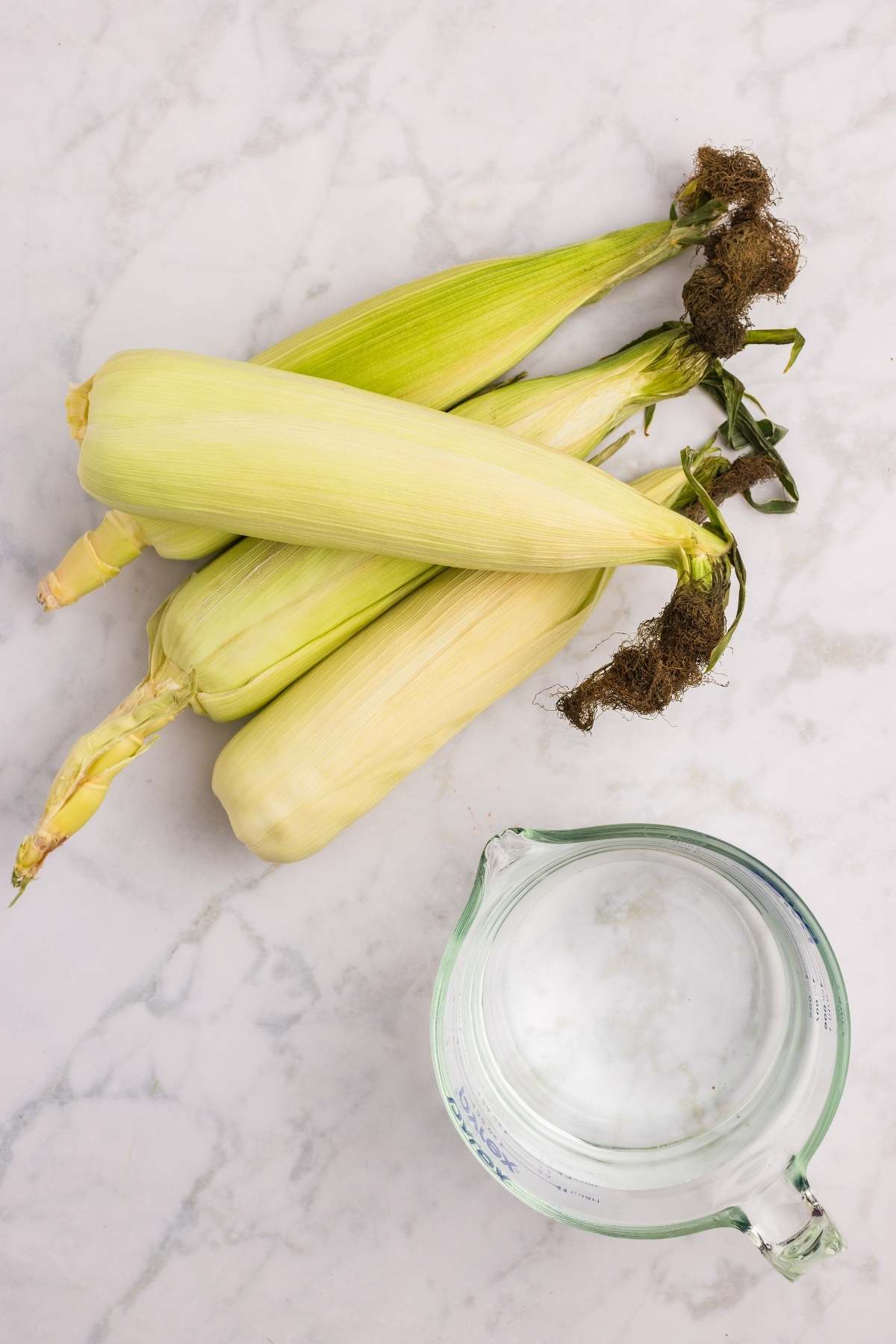 Four ears of corn with husks next to a glass measuring cup.
