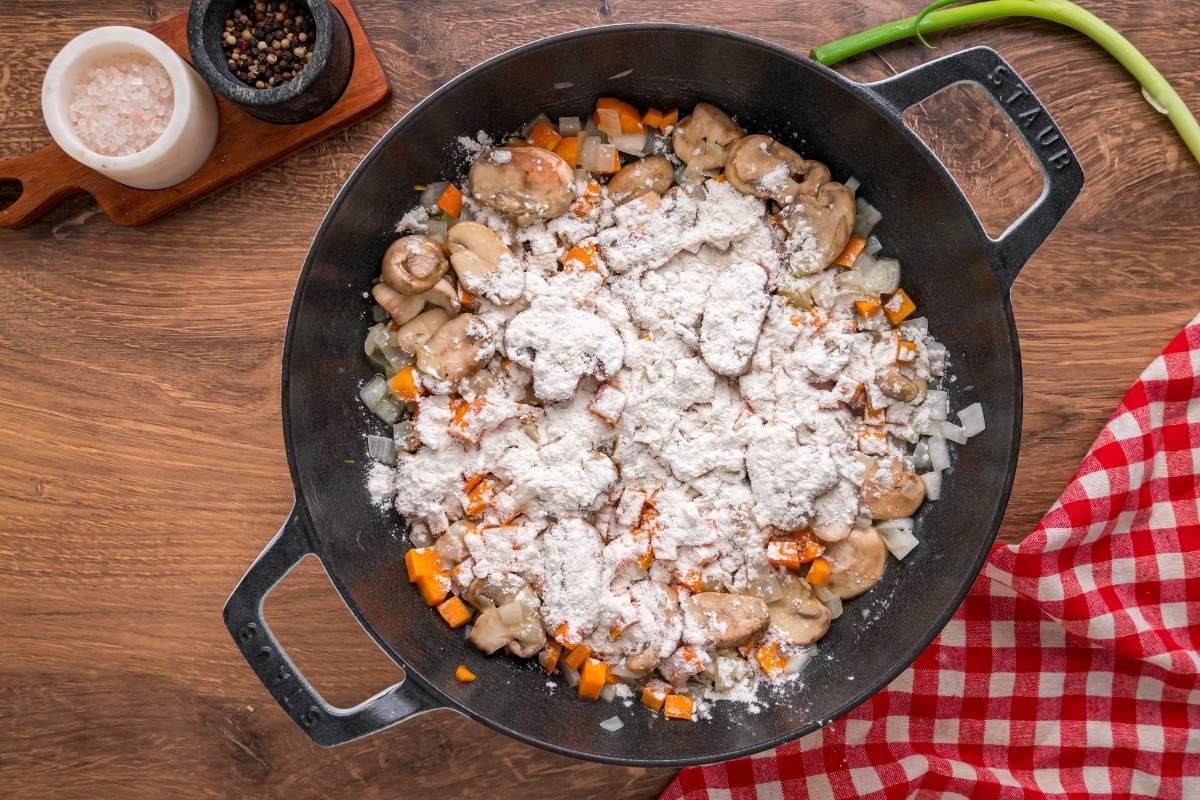 Flour sprinkled over cooked vegetables in black skillet.