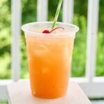 : Single glass of orange rum punch with cherry garnish and green striped straw.