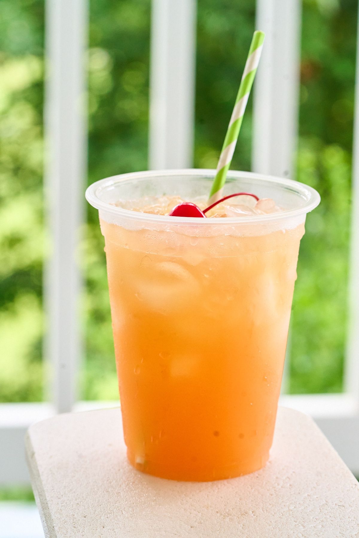 Glass of orange rum punch with cherry and green striped straw outdoors.