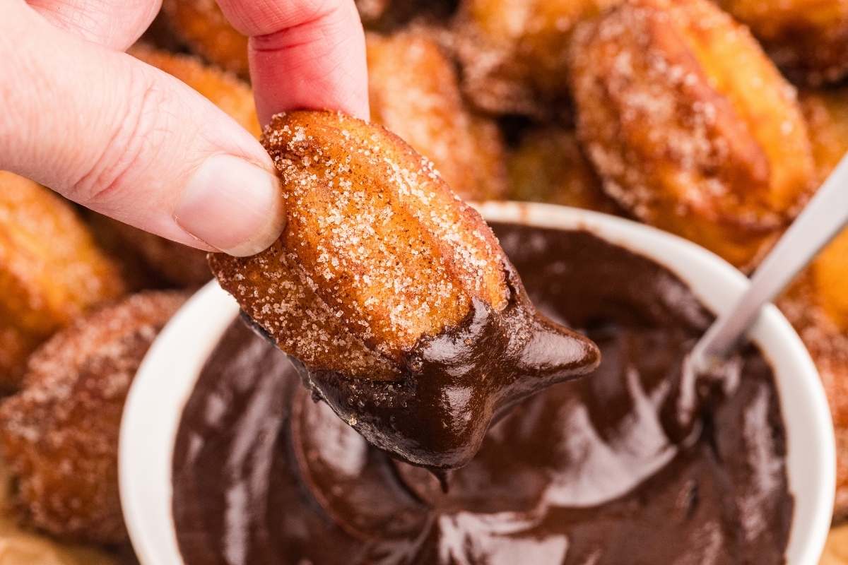 Hand holding cinnamon sugar churro bite dipped in chocolate sauce above bowl.