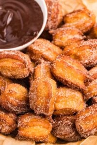 Cinnamon sugar coated churro bites arranged on parchment with bowl of chocolate sauce.