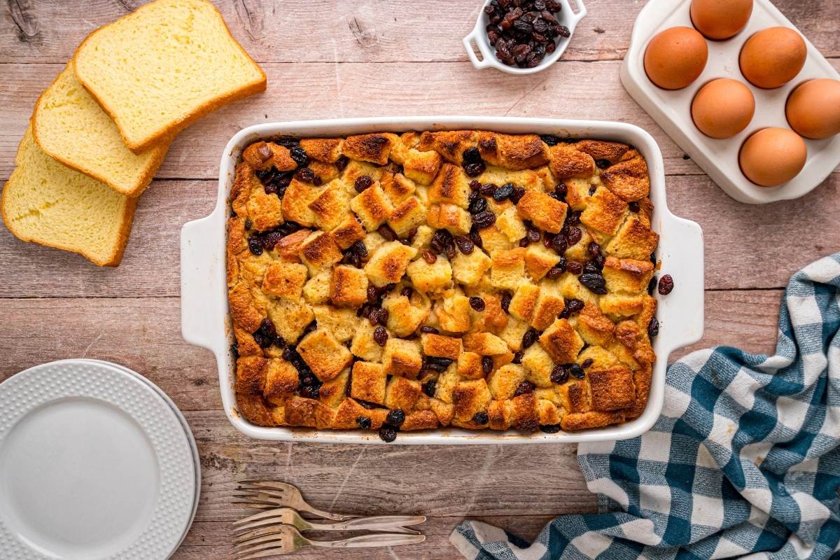 Baked bread pudding in white dish surrounded by ingredients on wood table.